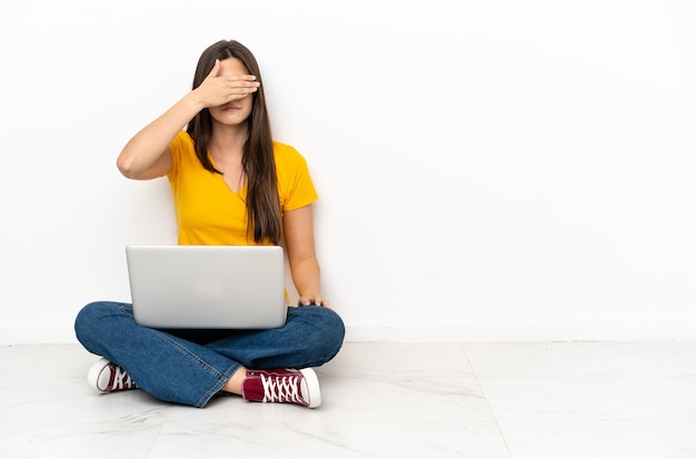 Jovem mulher com um laptop sentado no chão, cobrindo os olhos com as mãos. Não quero ver nada