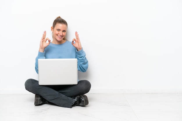 Jovem mulher com um laptop sentado no chão, mostrando um sinal de ok com os dedos