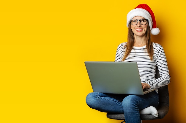 Jovem mulher com um laptop, usando um chapéu de Papai Noel e sentada em uma cadeira