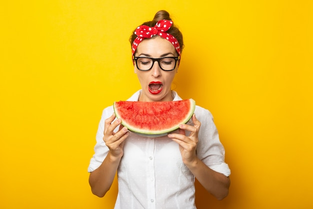 Jovem mulher com um sorriso em uma bandana vermelha e óculos come uma melancia em uma parede amarela.