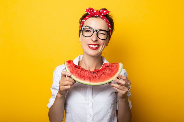 Jovem mulher com um sorriso em uma bandana vermelha e óculos come uma melancia em uma parede amarela.