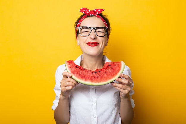 Jovem mulher com um sorriso em uma bandana vermelha e óculos come uma melancia em uma parede amarela.