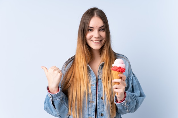 Jovem mulher com um sorvete de corneta na parede azul apontando para o lado para apresentar um produto