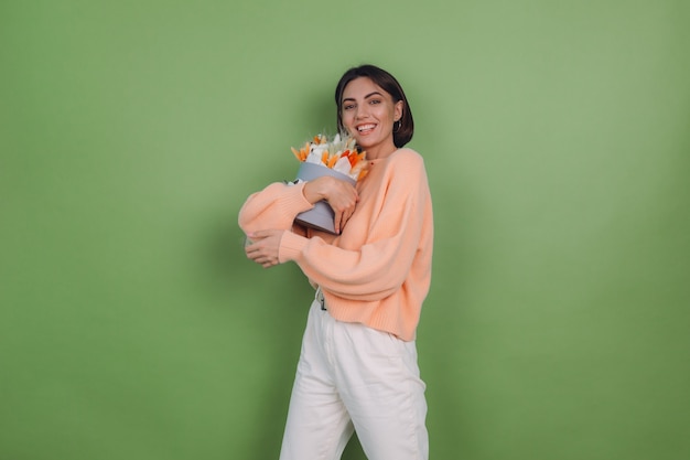 Jovem mulher com um suéter casual cor de pêssego isolado na parede verde-oliva segura uma caixa de flores brancas de laranja com flores de algodão, gypsophila, trigo e lagurus, para um presente feliz espantado surpreso