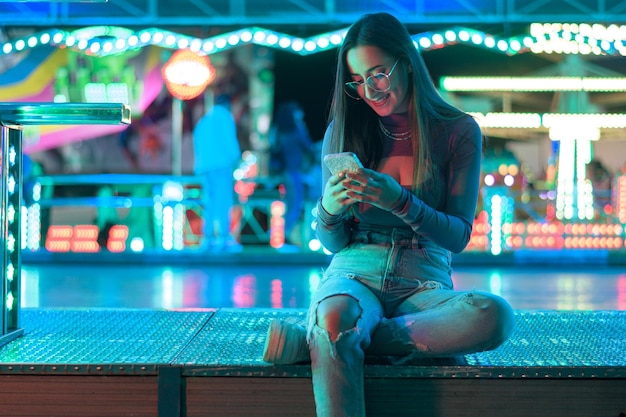 Jovem mulher com um telefone iluminado por luzes de parque de diversões