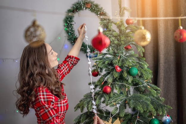 Jovem mulher com um vestido vermelho está decorando a árvore de Natal com brinquedos e bolas. Conceito de preparação para o Ano Novo, ambiente acolhedor e clima festivo.