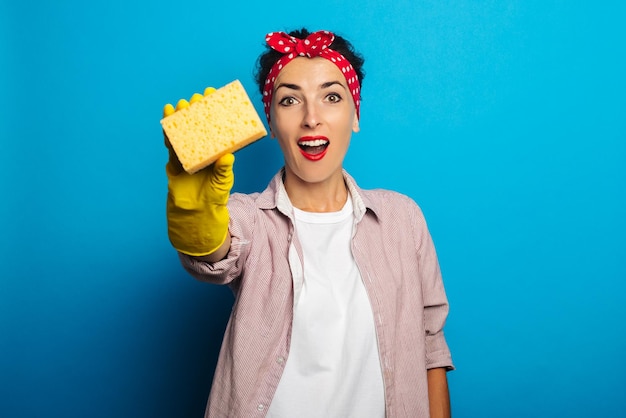 Jovem mulher com uma bandagem para o cabelo, em luvas amarelas, segurando uma esponja sobre um fundo azul.