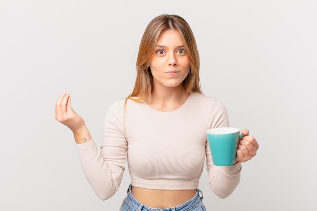 Jovem mulher com uma caneca de café fazendo um gesto de capice ou dinheiro, dizendo para você pagar