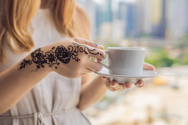 Jovem mulher com uma foto na mão de henna - mehendi bebe café pela manhã na varanda com vista para a cidade grande e os arranha-céus