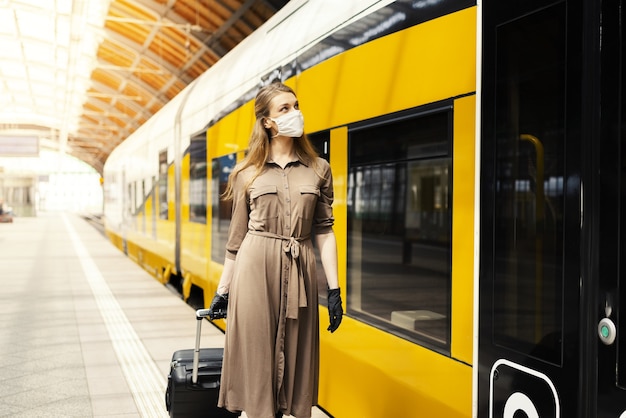 Jovem mulher com uma mala de luvas e uma máscara em uma estação de trem - COVID-19
