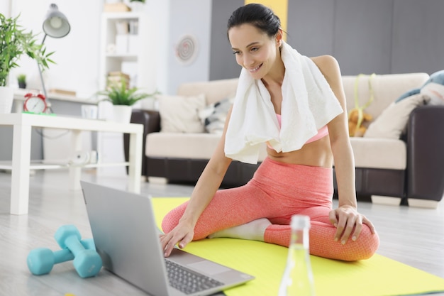 Jovem mulher com uma toalha em volta do pescoço, fazendo exercícios de esportes em casa na frente do laptop. Aptidão remota durante o conceito de pandemia COVID 19