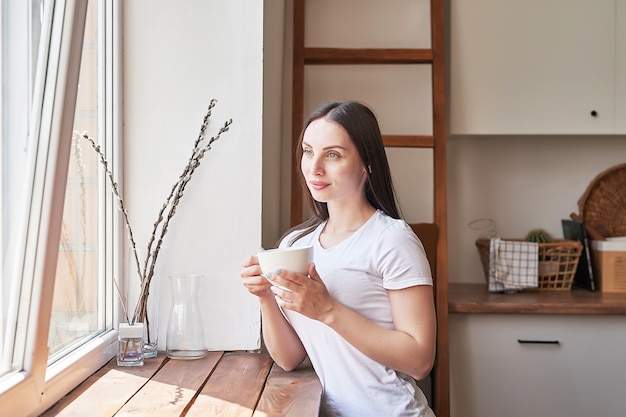 Jovem mulher com uma xícara de café ou chá na janela na cafeteria. Bom dia, conceito. Café da manhã aconchegante. Garota no café.