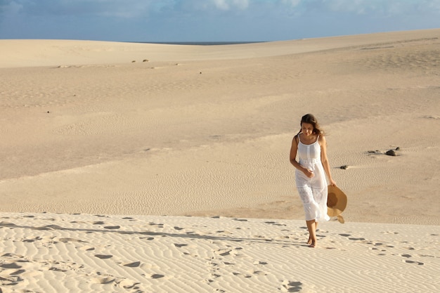 Jovem mulher com vestido branco e chapéu andando nas dunas do deserto solitárias