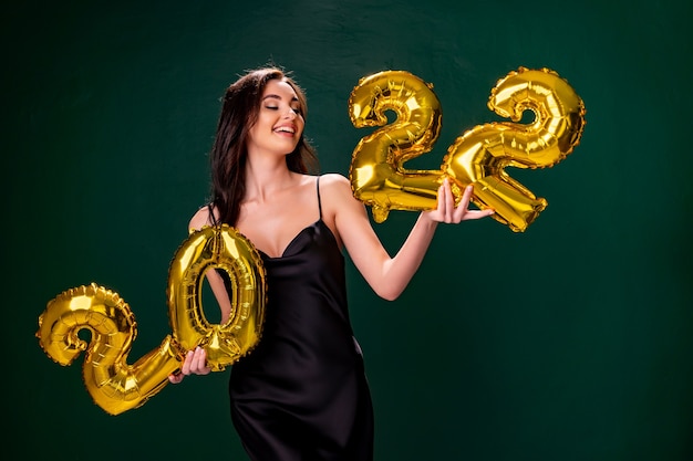 Jovem mulher com vestido de cocktail com maquiagem brilhante, comemorando o ano novo e segurando balões dourados.