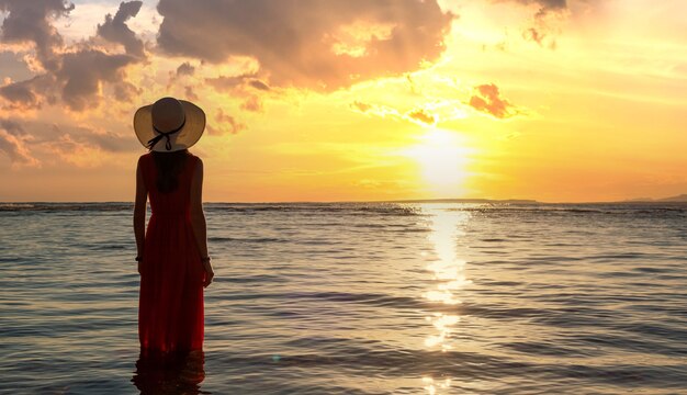 Jovem mulher com vestido longo vermelho e chapéu de palha em pé na água do mar na praia, apreciando a vista do sol nascente no início da manhã de verão.