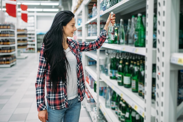 Jovem mulher comprando água mineral no supermercado. Cliente do sexo feminino em compras em hipermercado, departamento de bebidas