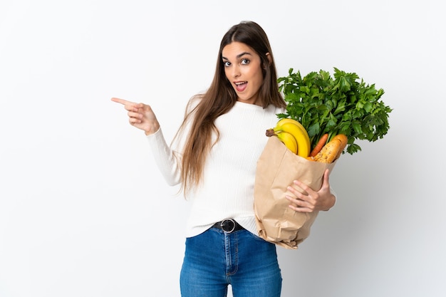 Jovem mulher comprando comida isolada em uma parede branca apontando o dedo para o lado e apresentando um produto