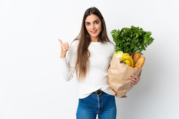 Jovem mulher comprando comida isolada em uma parede branca apontando para o lado para apresentar um produto