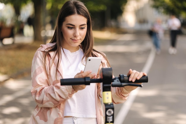 Foto jovem mulher conectando uma scooter elétrica ao ar livre, usando o telefone para pagamento sem contato