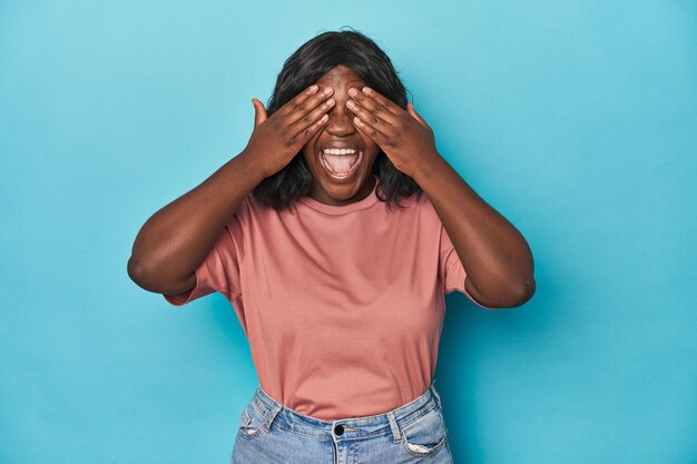 Jovem mulher curvilínea afro-americana cobre os olhos com as mãos e sorri amplamente esperando por uma surpresa