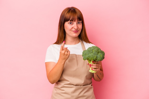 Jovem mulher curvilínea caucasiana, cozinhando em casa segurando brócolis isolado no fundo rosa, apontando com o dedo para você como se fosse um convite para se aproximar.
