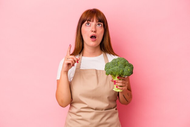 Jovem mulher curvilínea caucasiana cozinhando em casa segurando brócolis isolado no fundo rosa, apontando para cima com a boca aberta.
