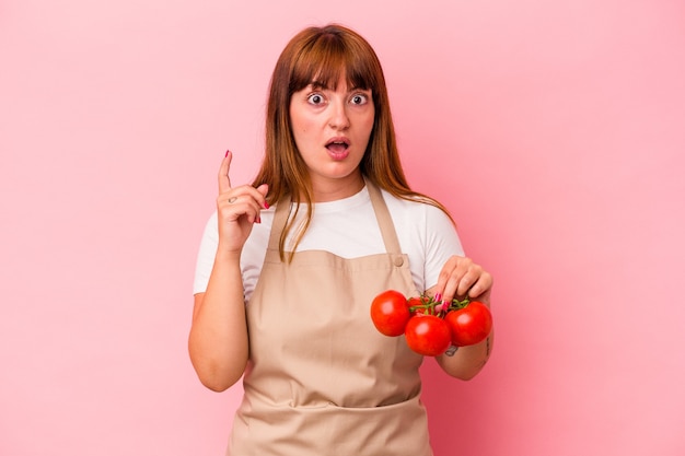 Jovem mulher curvilínea caucasiana cozinhando em casa segurando tomates isolados no fundo rosa, tendo uma ideia, o conceito de inspiração.