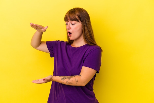 Jovem mulher curvilínea caucasiana isolada em fundo amarelo chocada e espantada, segurando um espaço de cópia entre as mãos.