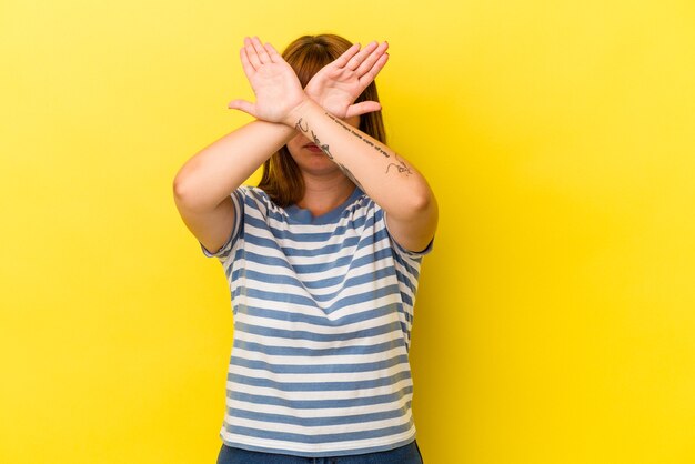 Jovem mulher curvilínea caucasiana isolada em fundo amarelo, mantendo os dois braços cruzados, conceito de negação.