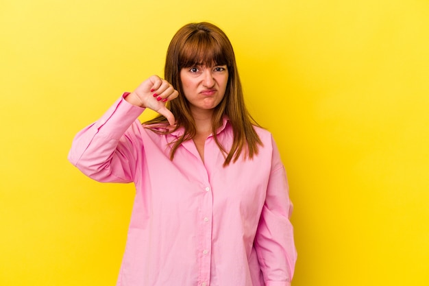 Jovem mulher curvilínea caucasiana isolada em fundo amarelo, mostrando o polegar para baixo e expressando antipatia.