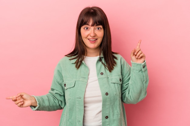 Jovem mulher curvilínea caucasiana isolada em fundo rosa, apontando para diferentes espaços de cópia, escolhendo um deles, mostrando com o dedo.