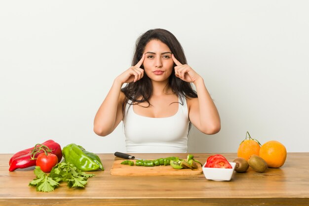 Jovem mulher curvilínea preparando uma refeição saudável focada em uma tarefa, mantendo os dedos apontando a cabeça.