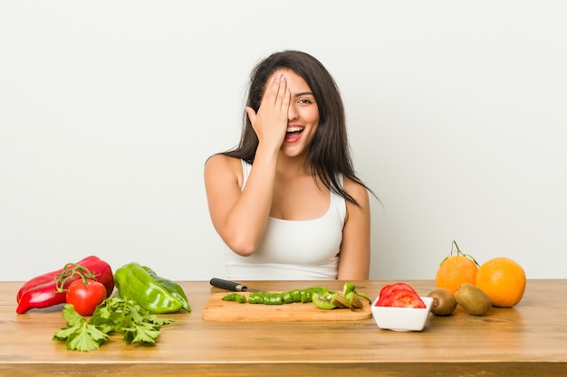 Jovem mulher curvilínea preparando uma refeição saudável, se divertindo, cobrindo metade do rosto com a palma da mão.