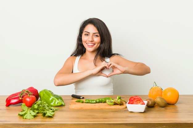 Jovem mulher curvilínea preparando uma refeição saudável, sorrindo e mostrando uma forma de coração com as mãos.