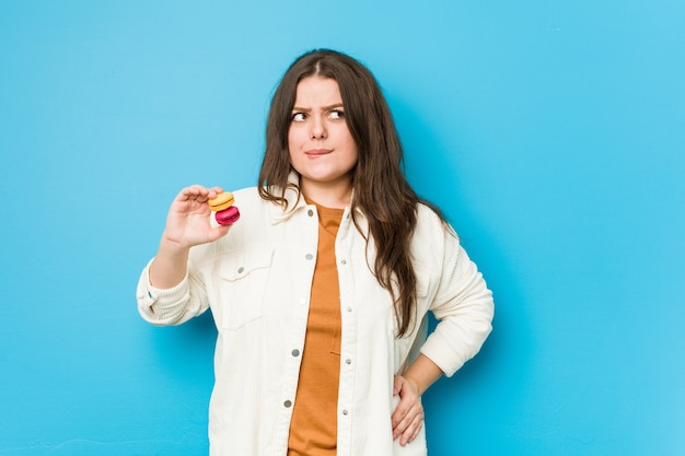 Jovem mulher curvilínea segurando macaroons confusa, sente-se duvidosa e insegura.