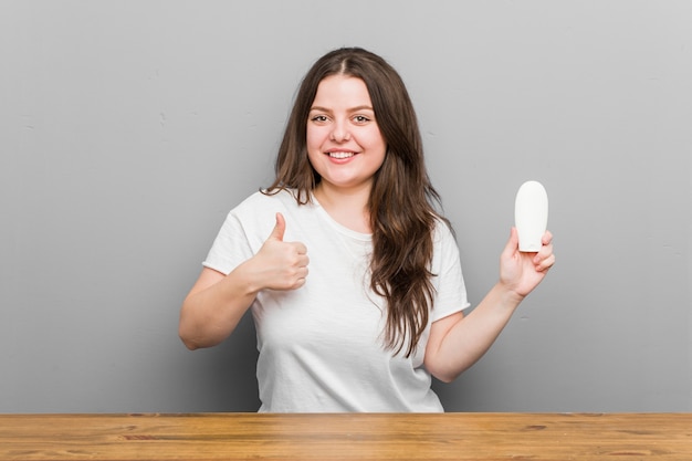 Jovem mulher curvilínea segurando um hidratante, sorrindo e levantando o polegar