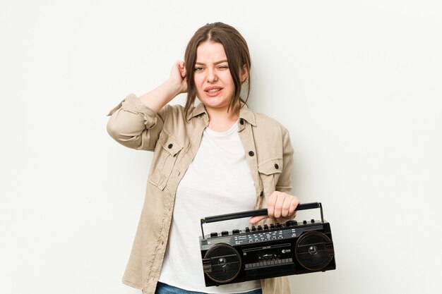 Jovem mulher curvilínea segurando um rádio retrô, cobrindo os ouvidos com as mãos.