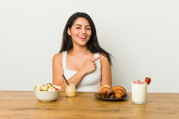 Jovem mulher curvilínea tomando café da manhã sorrindo e apontando para o lado, mostrando algo no espaço em branco.