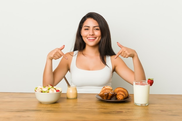 Jovem mulher curvilínea tomando um café da manhã aponta para baixo com os dedos, sentimento positivo.