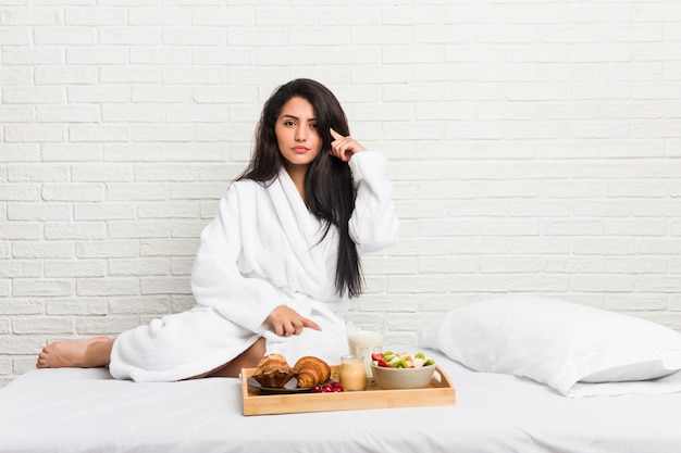 Jovem mulher curvilínea tomando um café da manhã na cama apontando o templo com o dedo, pensando, focado em uma tarefa.