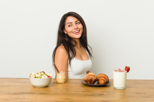 Jovem mulher curvilínea tomando um café da manhã parece de lado sorridente, alegre e agradável.