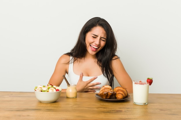 Jovem mulher curvilínea tomando um café da manhã ri alegremente e se diverte mantendo as mãos no estômago.