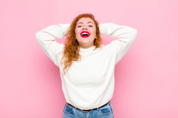 Jovem mulher de cabeça vermelha, olhando feliz, despreocupada, amigável e relaxada, curtindo a vida e o sucesso, com uma atitude positiva sobre a parede rosa