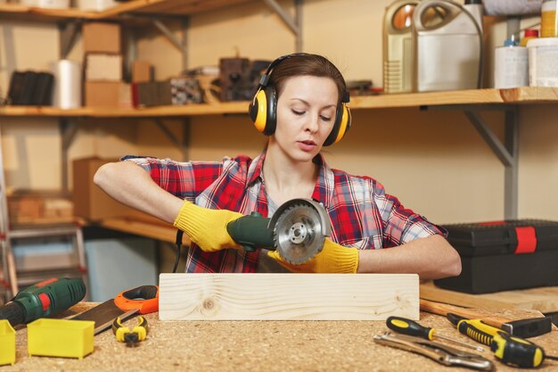 Jovem mulher de cabelo castanho com camisa xadrez, camiseta cinza, fones de ouvido com isolamento acústico, luvas amarelas trabalhando em uma oficina de carpintaria em uma mesa de madeira com ferramentas diferentes, serrando madeira com serra elétrica