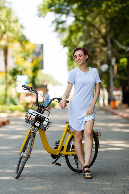 Jovem mulher de cabelo rosa caminha no parque com uma bicicleta, tomando sorvete no verão. Meio de transporte ambiental