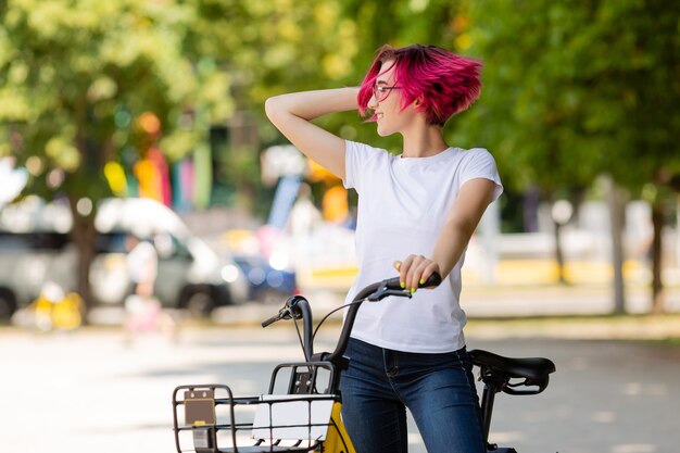 Jovem mulher de cabelo rosa caminha no parque com uma bicicleta, tomando sorvete no verão.