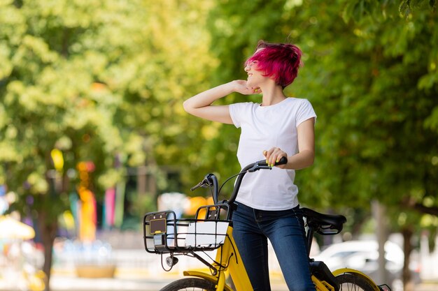 Jovem mulher de cabelo rosa caminha no parque com uma bicicleta, tomando sorvete no verão.