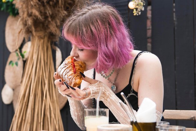 Foto jovem mulher de cabelo rosa sentado no café de rua ao ar livre comendo croissant