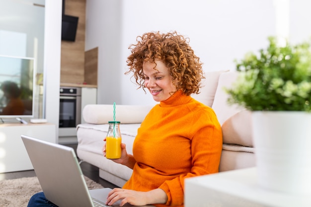 Jovem mulher de cabelo vermelho sorridente trabalhando em casa, ela está se conectando com um laptop e bebendo suco de laranja saudável. Linda garota ruiva navegando na web em seu laptop, conversando com seus amigos por videochamada