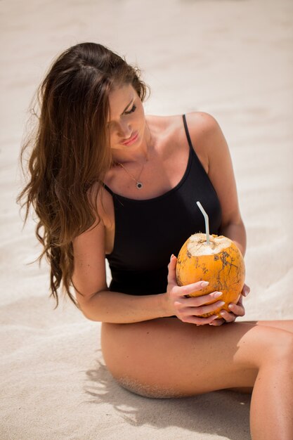 Foto jovem mulher de maiô preto com coquetel de coco na praia